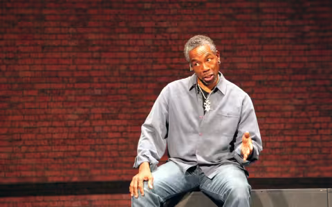 actor gesturing with his right hand sits in front of a brick wall