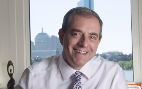 David Autor smiles in a white shirt and tie while seated in front of a winder. Through the window are buildings, trees, a body of water. 