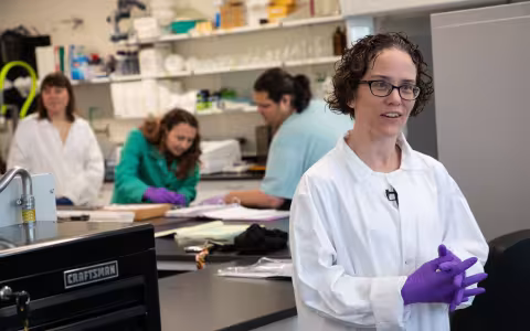 A woman with short brown curly hair and glasses wears a white lab coat and purple latex gloves, her hands clasped together as she smiles and speaks to someone outside the frame.