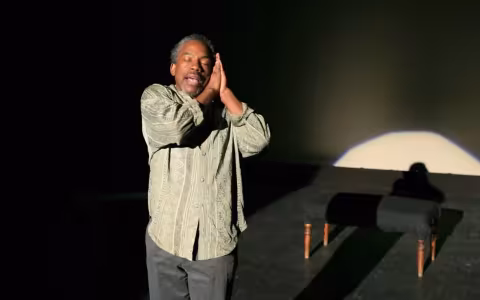 actor in light green shirt stands with eyes closed and hands clasped together on the side of his head