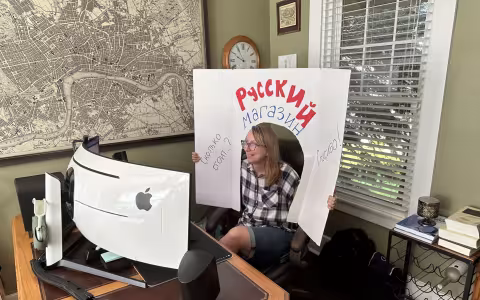 A woman, Maria Goebert, is seated in front of a computer screen in an office. She smiles and holds a white cardboard cutout featuring Russian phrases drawn on in marker over her head.