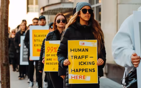 A line of people march, holding yellow anti-trafficking signs. At front is a women in sunglasses with a sign reading "Human Trafficking Happens Here"