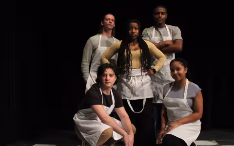 five actos in aprons stand or squat in a clump looking forward