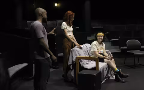 Three people appear in frame. A man stands to one side gesturing to a woman who is seated, as another woman stands directly behind her watching.