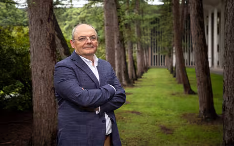 Andrei Lapenas stands in a suit with his arms folded, surrounded by trees, in front of the Academic Podium courtyard on the Uptown Campus.