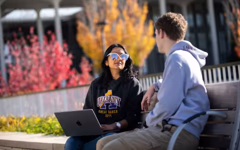 Two students wearing sweatshirts sit on a bench on a sunny fall day. One holds a laptop on their lap.
