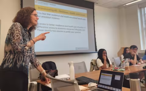 A woman in glasses stands in front of a screen with projected words about "residence, health and Causes" as students listen to her speak