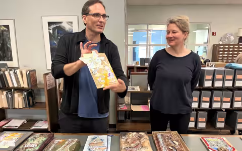 A man with short black hair and glasses holds a handmade art book while a woman with gray hair watches. They stand in front of a table topped with handmade books.