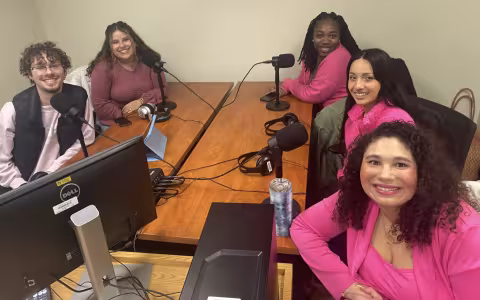 Five young people dressed in pink sit around a table topped with microphones, headphones and a Dell computer. They smile at the camera. 