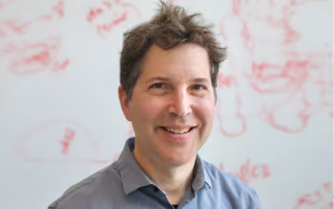 David Baker stands and smiles in front of an out-of-focus white board with red writing on it.