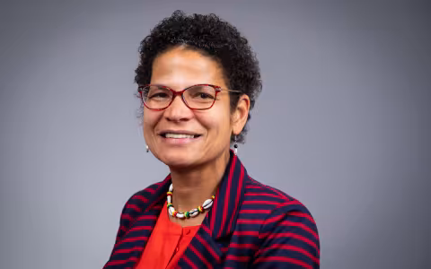 Portrait of Elizabeth Vásquez against a gray background. Vásquez is smiling, wearing red rimmed glasses, a shell necklace, red top and red and navy striped blazer.
