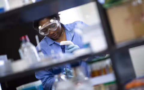 A student in a blue lab coat wearing gogles works with a pipette behind a bench in a laboratory with several beakers filled with liquids of various colors.