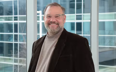 Portrait of Scott Tenenbaum, who is wearing clear-rimmed glasses, a dark brown blazer and tan turtleneck sweater. Tenenbaum is smiling, standing in front of a wall of glass windows.