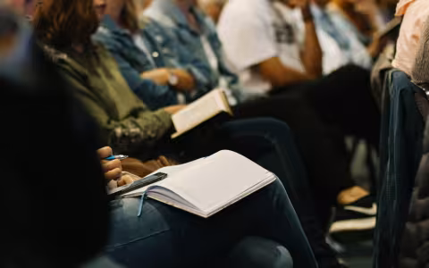 Closeup of seated people crowded in rows, with open notepads and books on their laps.