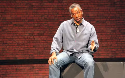 A seated man in jeans and a gray shirt gestures while speaking, his head tilted to one side.