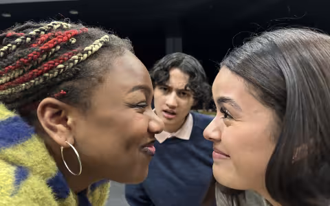 two actors in profile smile face to face with a third actor between them in the background