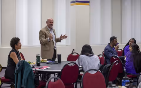 Paul Morgan stands at a table addressing a room of students gathered for a lunch meeting. Morgan is smiling, wearing a tan blazer and white button up shirt.