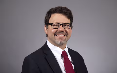 Portrait of a smiling man with short brown hair and glasses, wearing a suit and tie against a gray backdrop.