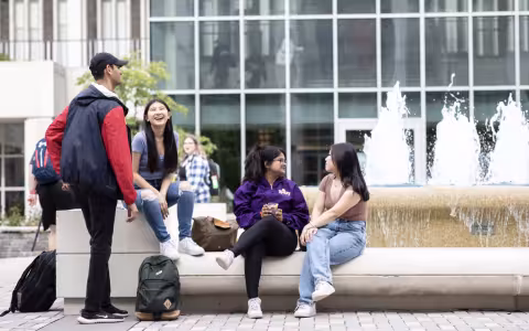 Students are seated and standing around a fountain on the UAlbany campus on a clear bright day.