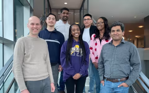 Six people pose together for a group portrait on an open stairwell in the RNA Institute atrium. There are five men and two women. All are smiling.