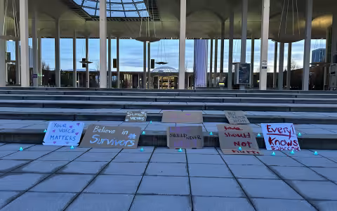 Image depicts eight painted signs made of cardboard, propped up on the front steps of UAlbany’s academic podium. The photo was taken in the evening, and the sky in the background is blue, lightly clouded and darkening. The signs say: your voice matters, believe in survivors, support victims, we love survivors, love should never hurt, love should not hurt, no means no, and everyone knows someone. Small green LED tea lights are scattered around the signs. 