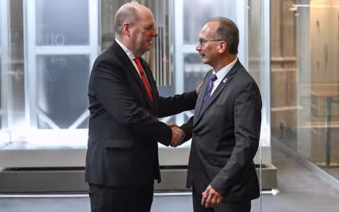 Two men in dark suits face each other and shake hands in front of a brightly illuminated enclosure holding a quantum computer.