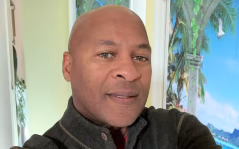 Superintendent James poses in front of a wall with a painting of a palm tree and blue sky. James is smiling and is wearing a dark gray mock turtleneck sweater with a wooden button at the neck. 