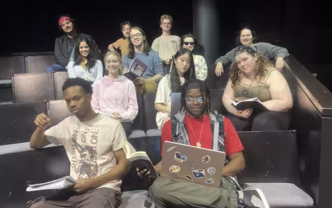 A dozen young people sit in auditorium seats with laptops, notebooks and books and pose for a photo.