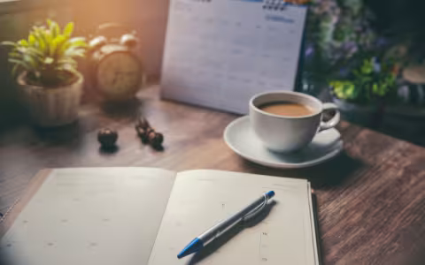A study desk with a clock, planner, cup of coffee and plants on it. 