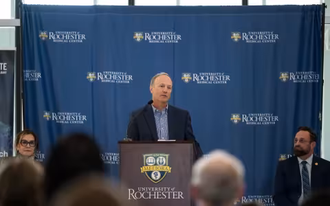 A man in a jacket stands a brown University of Rochester lectern facing an audience. 