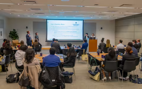 A room full of college-age students and business mentors sit at tables and view a projection screen