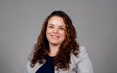 Sarah Domoff poses for a portrait against a gray background wearing a light gray jacket over a navy blue shirt. Domoff is smiling and has brown curly hair. 