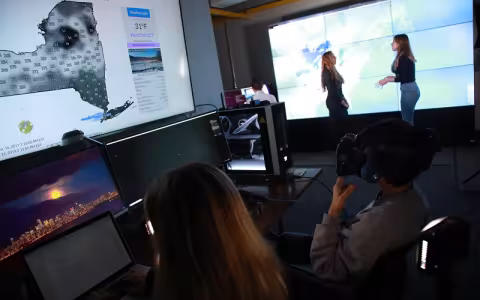 Two people sit a screens in a darkened computer lab, with one of them wearing VR goggles, as to people speak in the background.
