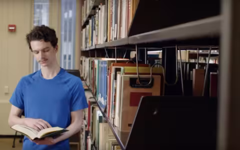 A young man in a blue shirt holds a book open as he stands next to library shelves.