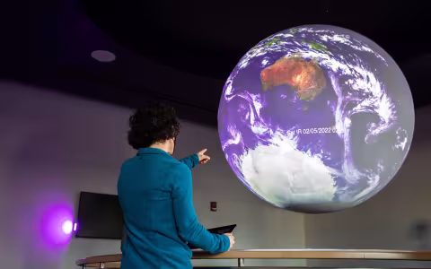 DAES Assistant Professor Aubrey Hillman points at a map of Earth's atmospheric conditions inside the Science on a Sphere room.
