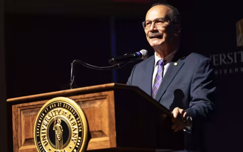 UAlbany President Havidán Rodríguez stands behind a wooden podium with the UAlbany seal and speaks into a microphone.