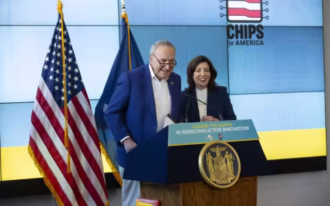 A man in a blue sport jacket and a woman in a dark business suit stand at a lectern next to an American flag. The sign on the lectern says: "Leading the Nation in Semiconductor Innovation"