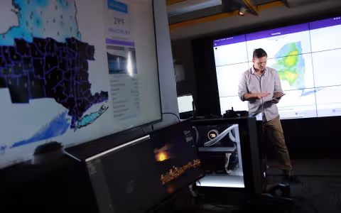 A man with an ipad looks down while a weather map of NEw York state appears in the foreground at UAlbany's XCite Lab.