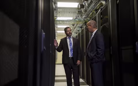 Two men in dark suits stand facing one another between two long rows of computer racks.