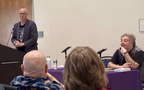 Dr. Ian Singleton stands up at a lectern, while Russian author Dmitry Bykov sits at a table in front of a microphone.