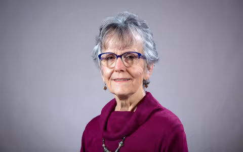 A woman with short gray hair and glasses wears a pink sweater and poses for a portrait against a gray backdrop