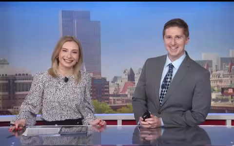 UAlbany alum Reid Kisselback and a female colleague smile and pose while sitting behind a newscasters desk. The Albany skyline is projected behind them.