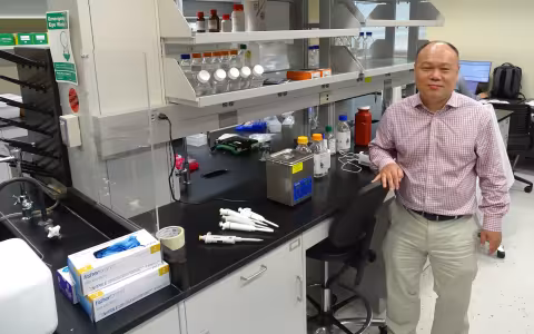 A man wearing a plaid button-down shirt and khaki pants poses, smiling, next to a lab bench covered in various pieces of chemistry equipment including pipettes, latex gloves, and plastic bottles containing clear liquids.