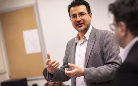 A man with short black hair and glasses in a gray blazer is pictured in a classroom.