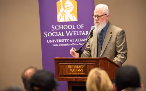 Eric Hardiman behind a podium. Behind him is a purple banner that says "School of Social Welfare". 