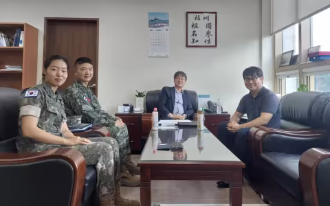 Two military personnel wearing uniforms are seated next to two civilians in an office. They smile and pose for a photo.