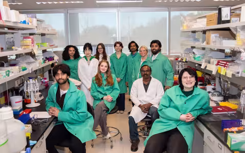  Eleven people wearing lab coats gather for a group photo in a brightly lit lab. They are flanked by lab benches and shelving packed with lab supplies and equipment. 