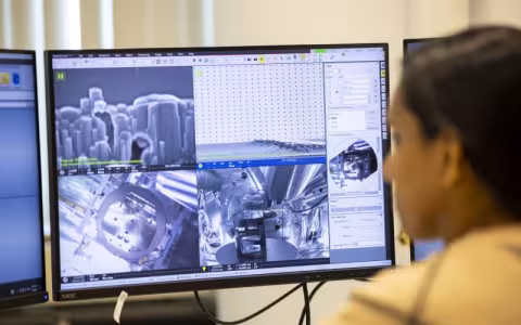 A woman's face is blurred in the foreground as she looks at computer screens with gray images that show heavily magnified computer chip designs. 