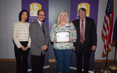 Four people stand in front of UAlbany banners. One is holding her Milestone Ceremony award.