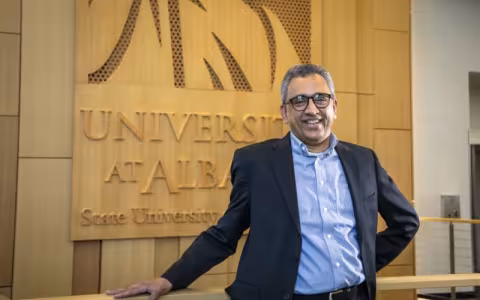A man in a dark suit coat and button-down shirt smiles in front of a wood-paneled wall with the words "University at Albany."